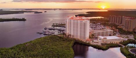 Un hotel con varias construcciones, un puerto deportivo con barcos y un atardecer sobre el agua.