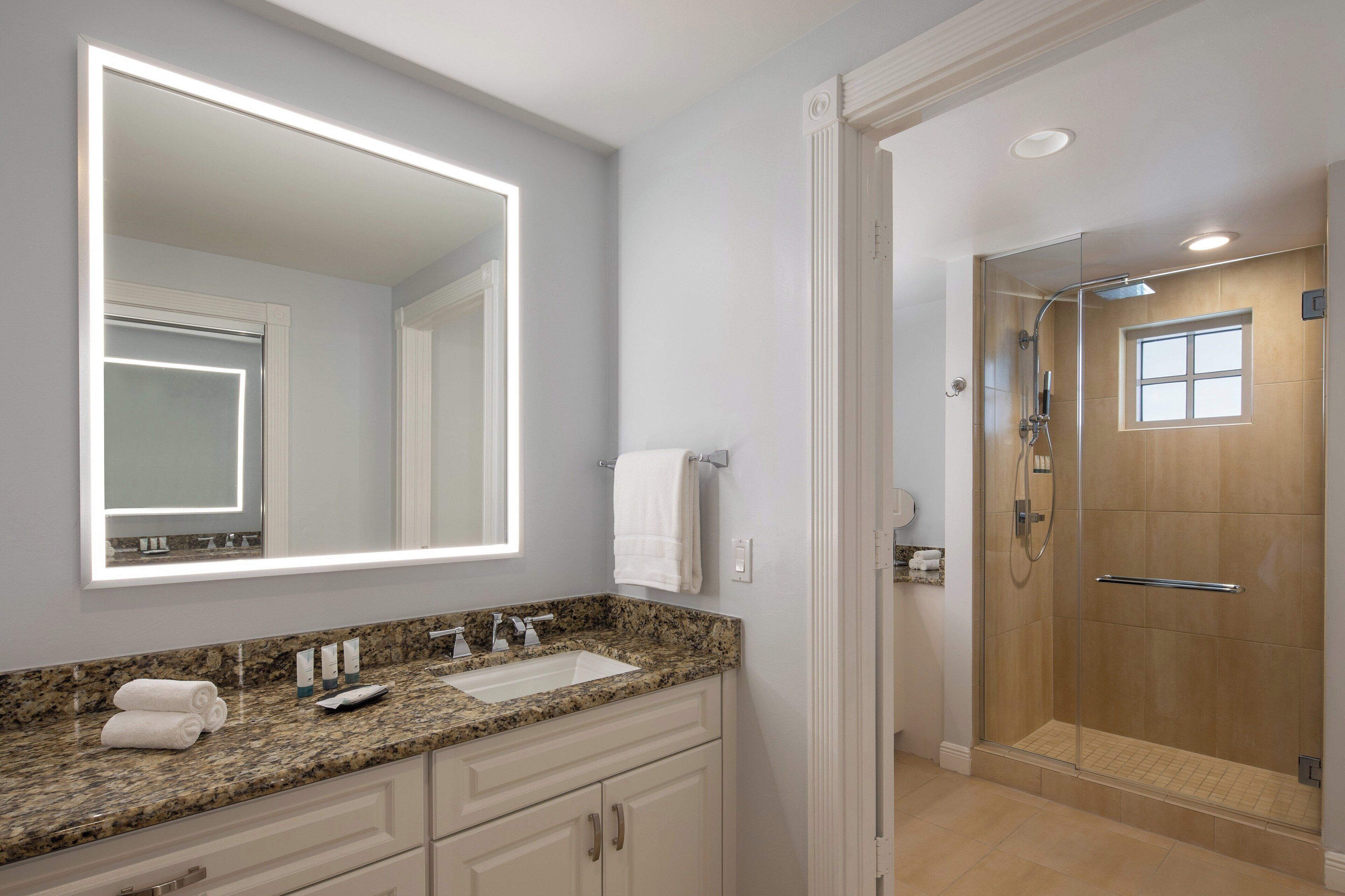 A bathroom with a granite countertop, a large mirror, a glass-enclosed shower, and a white towel on the wall.