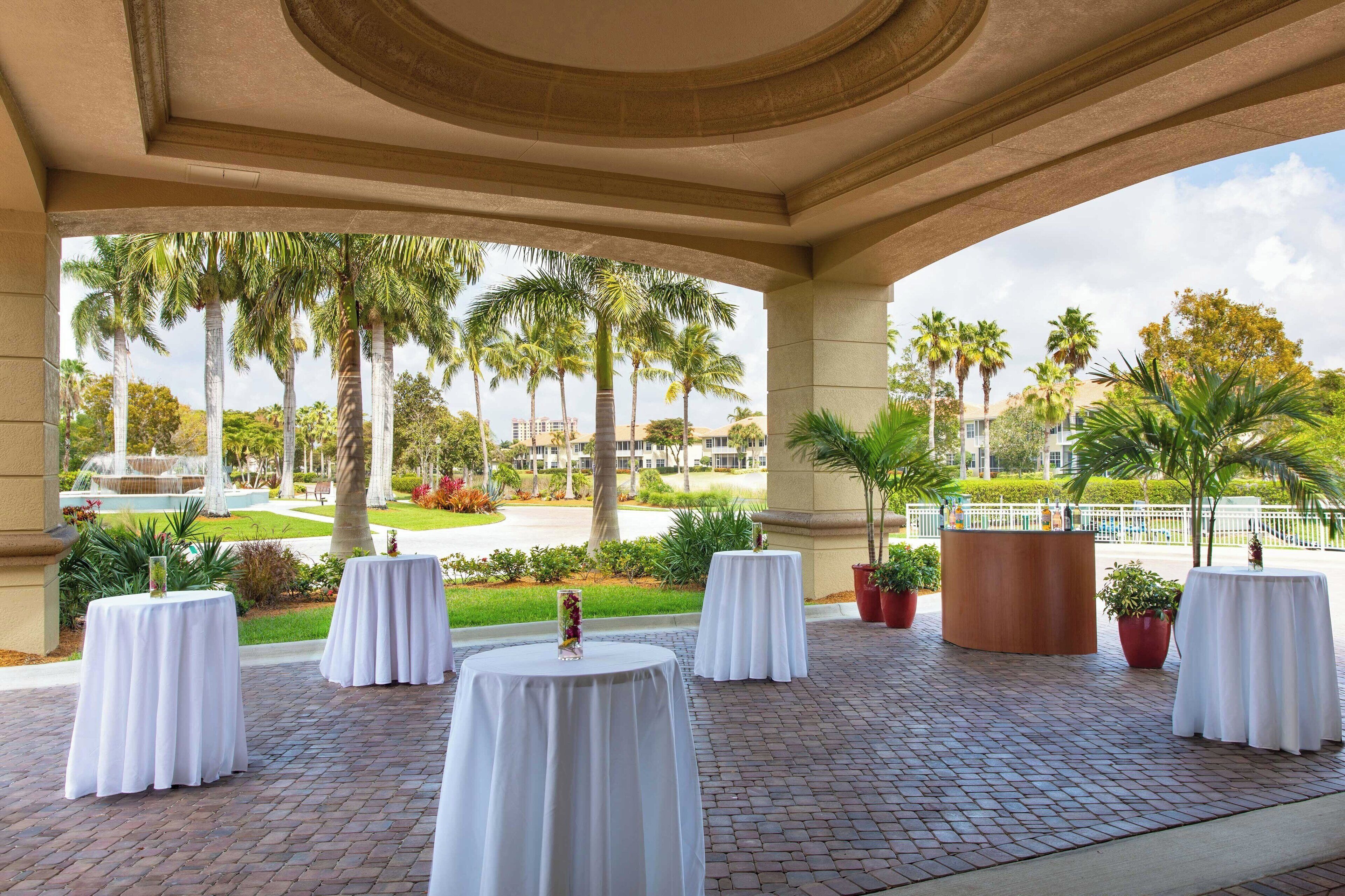 A covered outdoor area with white tables, palm trees, and a brick floor.