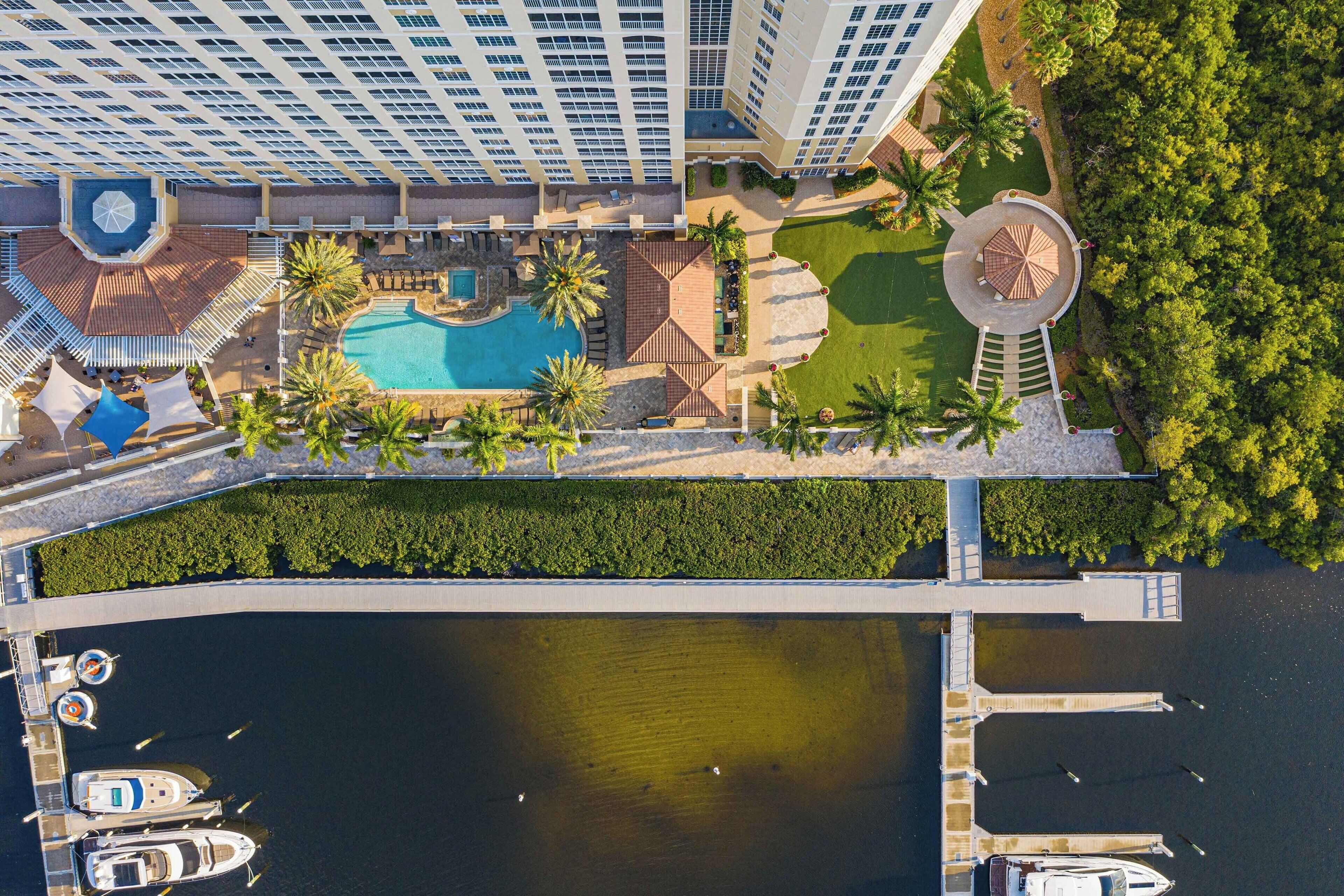 An aerial view of a hotel with a swimming pool, palm trees, and a dock with boats.