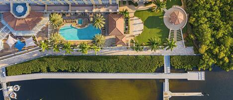 An aerial view of a hotel with a swimming pool, palm trees, and a dock with boats.