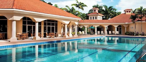 Une piscine à l’eau d’un bleu cristallin, entourée de chaises longues et de parasols, et un bâtiment surmonté d’un toit en tuiles.