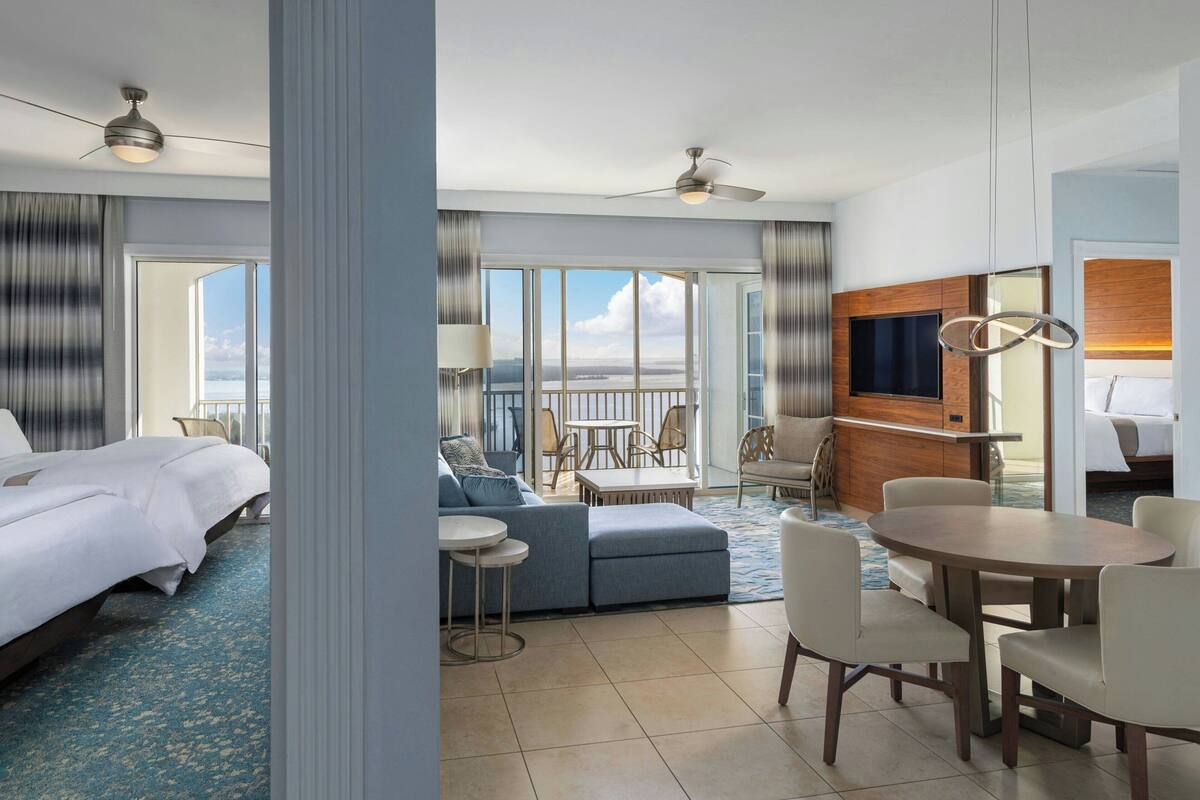 Quarto de hotel com cama, sofá, mesa de jantar com cadeiras, televisão e varanda com vista.