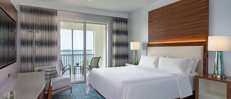 A hotel room with a bed, a desk, a chair, a balcony with a view, and a ceiling fan.