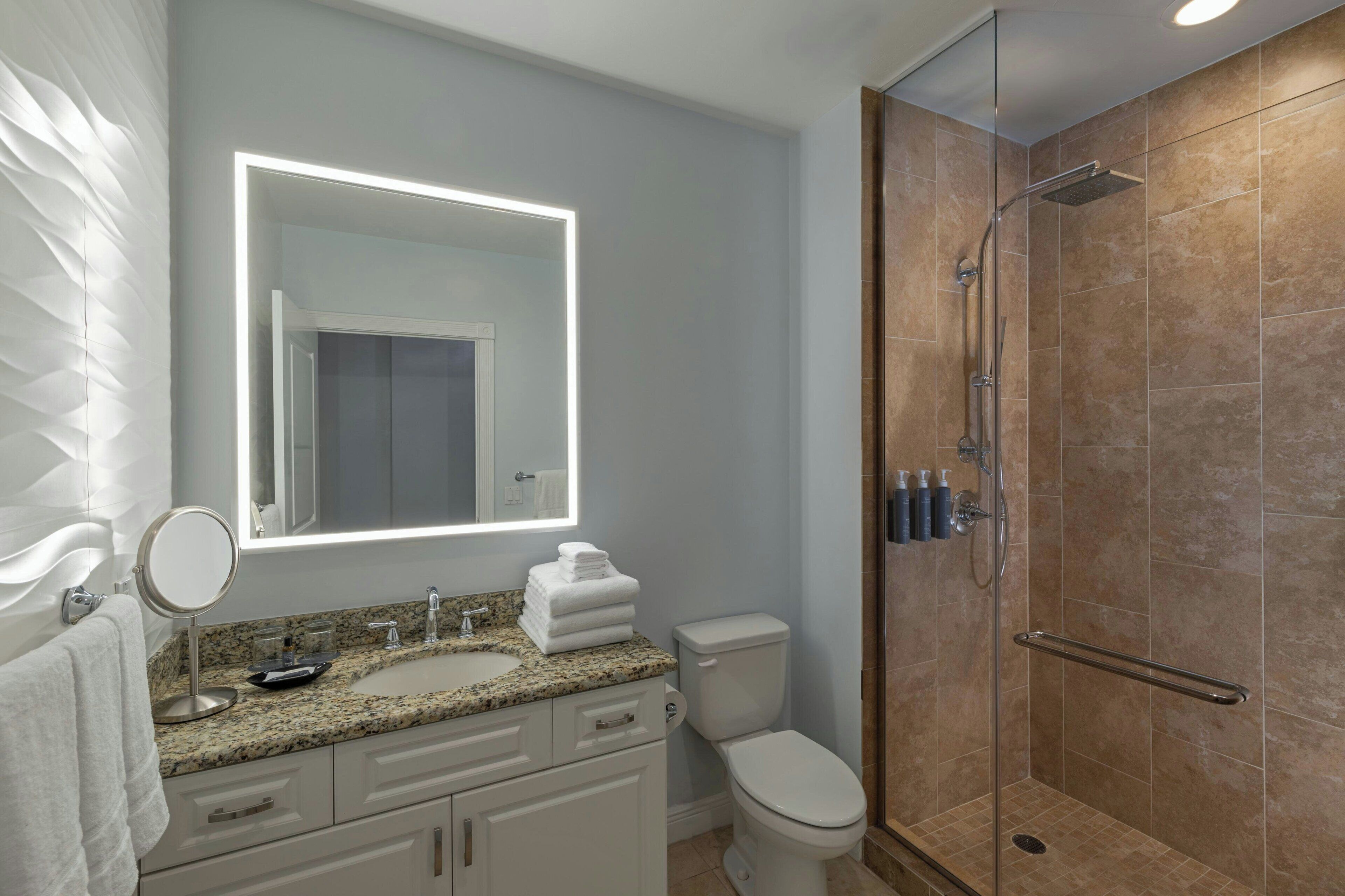 A modern bathroom with a granite countertop, a round mirror, a toilet, and a glass-enclosed shower.