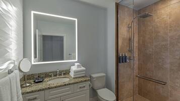 A modern bathroom with a granite countertop, a round mirror, a toilet, and a glass-enclosed shower.
