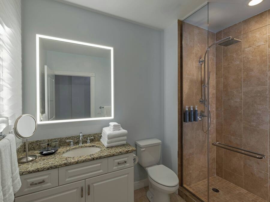 A modern bathroom with a granite countertop, a round mirror, a toilet, and a glass-enclosed shower.