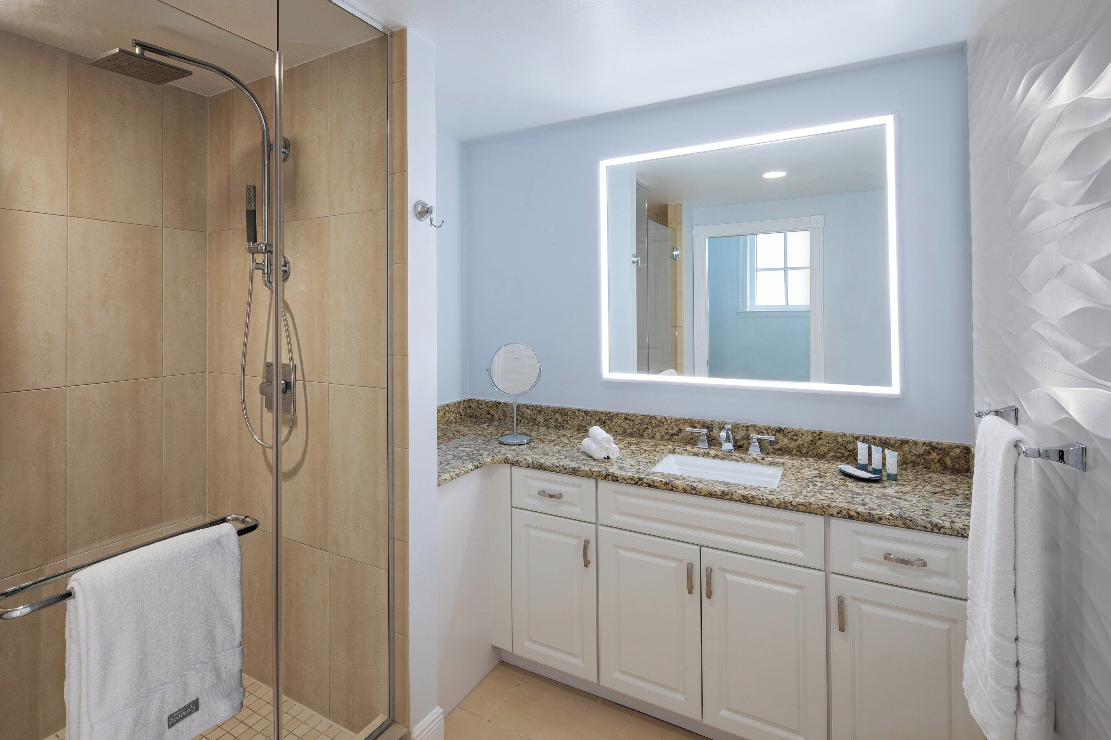 A modern bathroom with a glass shower enclosure, a granite countertop sink, and a large mirror with integrated lighting.