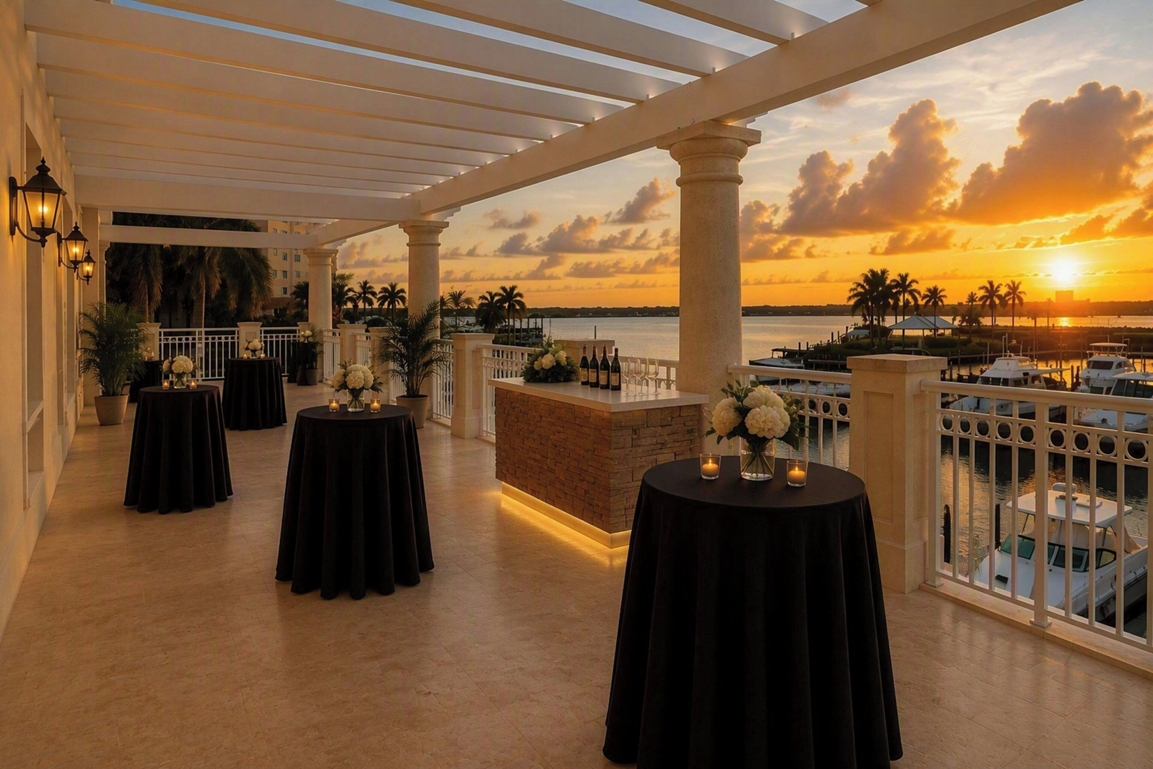 An outdoor terrace with tables set for dining, overlooking a marina and sunset.