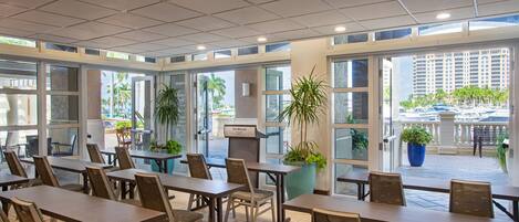 A spacious indoor dining area with multiple tables and chairs, large windows, and potted plants.