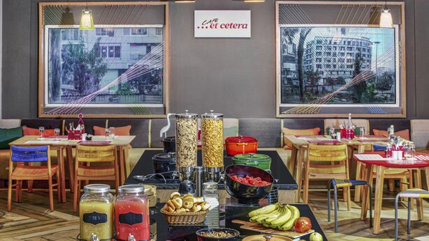 A breakfast buffet with various food items and beverages, tables with chairs, and two large windows with city views. - Ginger Bangalore Whitefield (Bengaluru)
