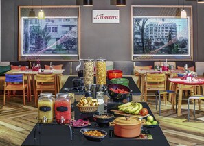 Un bufé de desayuno con diversas opciones de comida y bebidas, mesas con sillas y dos ventanas amplias con vistas a la ciudad.