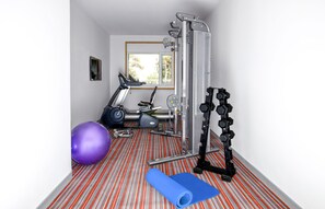 A small, brightly lit room with a red and white striped carpet, a stationary bike, a purple exercise ball, a blue yoga mat, and a weightlifting bench.