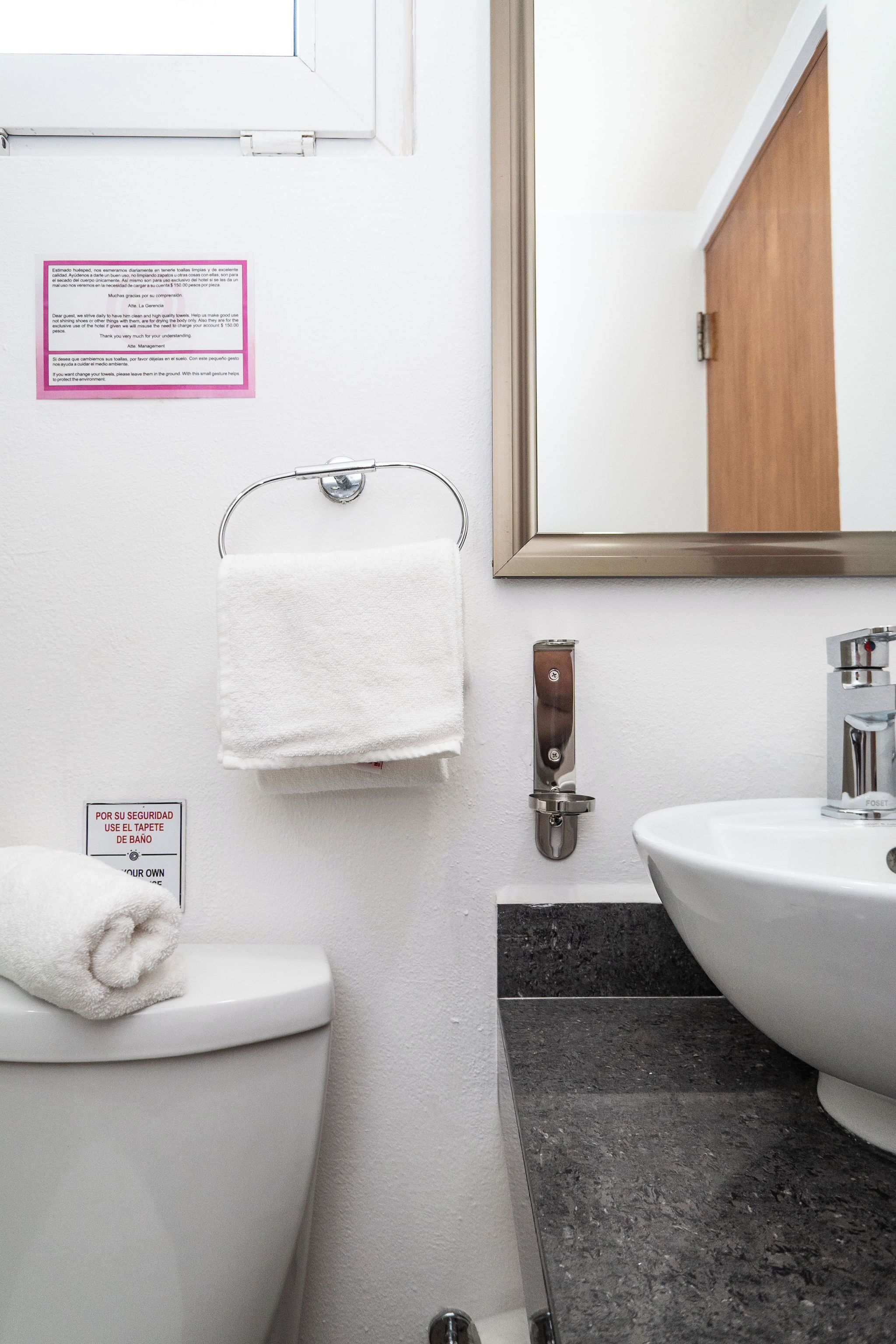 A bathroom with a white toilet, a sink, and a mirror. There is a towel hanging on a rack, and a sign on the wall.