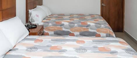A hotel room with a wooden headboard, a bed with a patterned bedspread, a bedside table with a lamp, and an air conditioning unit on the wall.