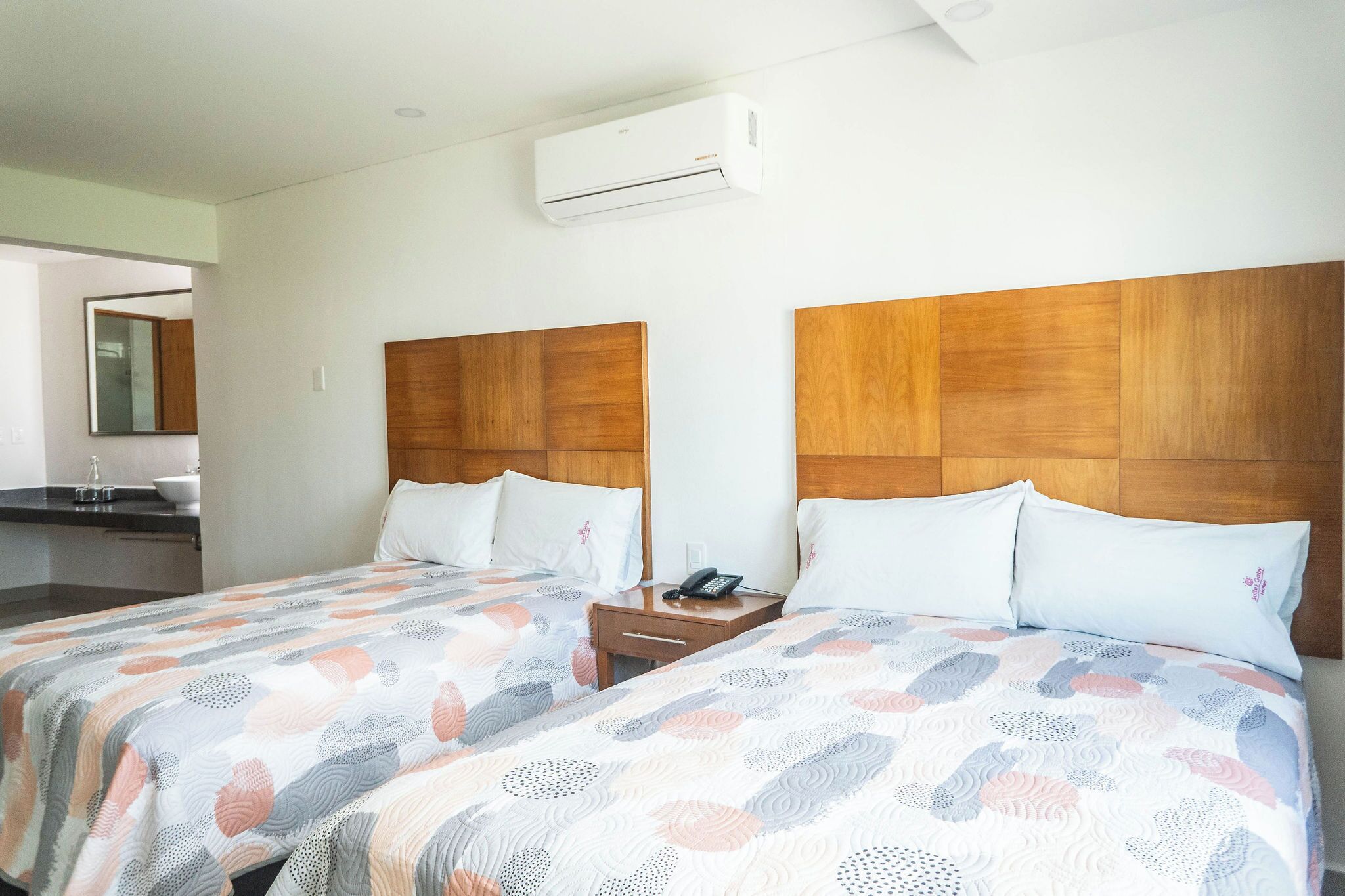 Une chambre d’hôtel avec deux lits, des têtes de lit en bois, une table de chevet avec un téléphone et un appareil de climatisation fixé au mur.