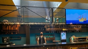 A bar area with a bartender, wooden bar counter, stools, and a television screen displaying a ship. - Tunisie Confort (Tunis)