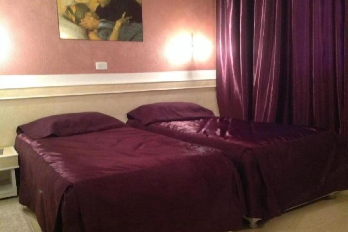 A room with two beds covered in purple bedding, a wall-mounted light, and a painting above the beds.