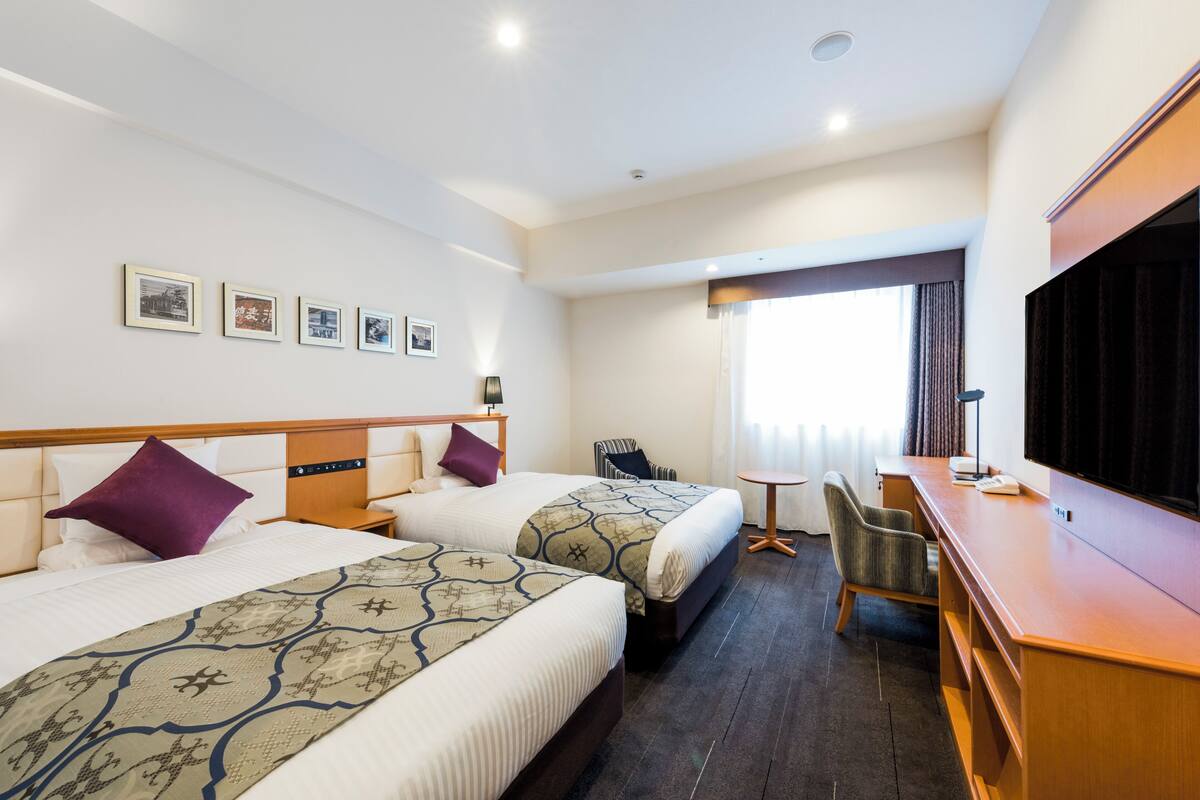 Ein Hotelzimmer mit zwei Betten, einem Schreibtisch, einem Sessel, einem Fernseher und gerahmten Bildern an der Wand.