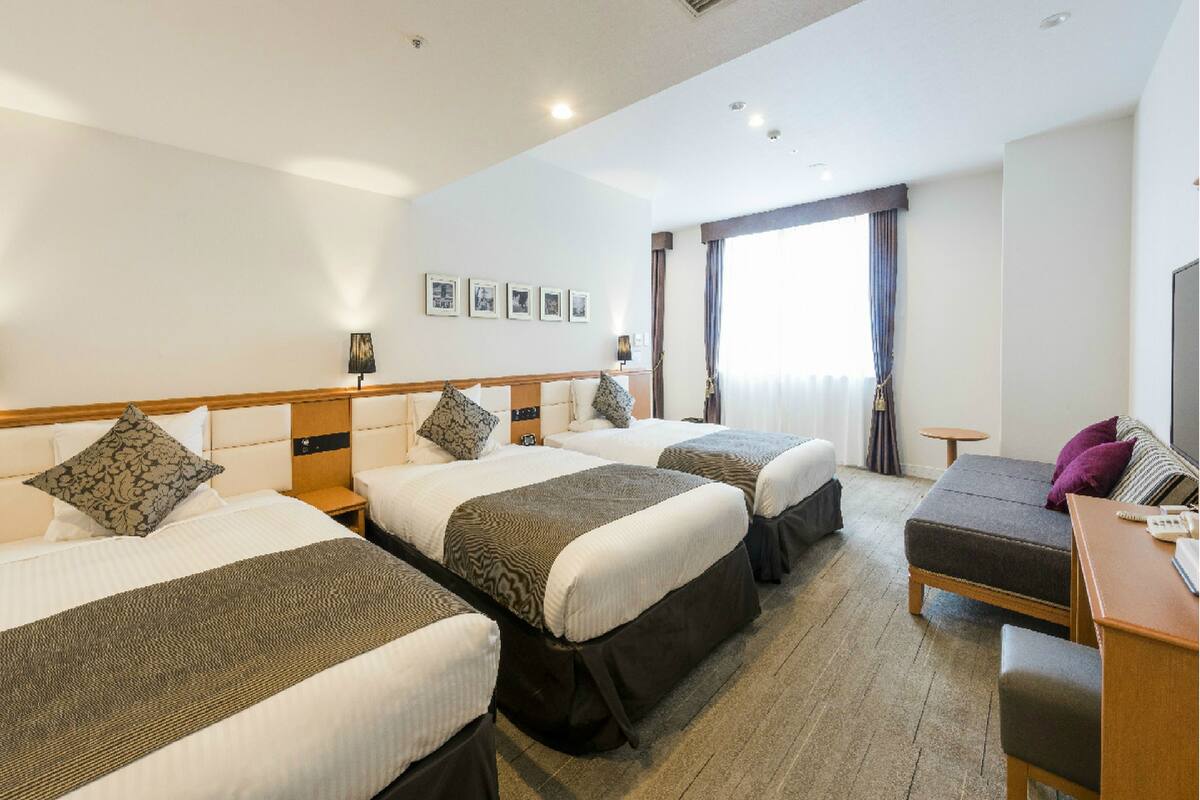 Ein Hotelzimmer mit drei Betten, einem Schreibtisch, einem Sessel und einem großen Fenster mit Vorhängen.