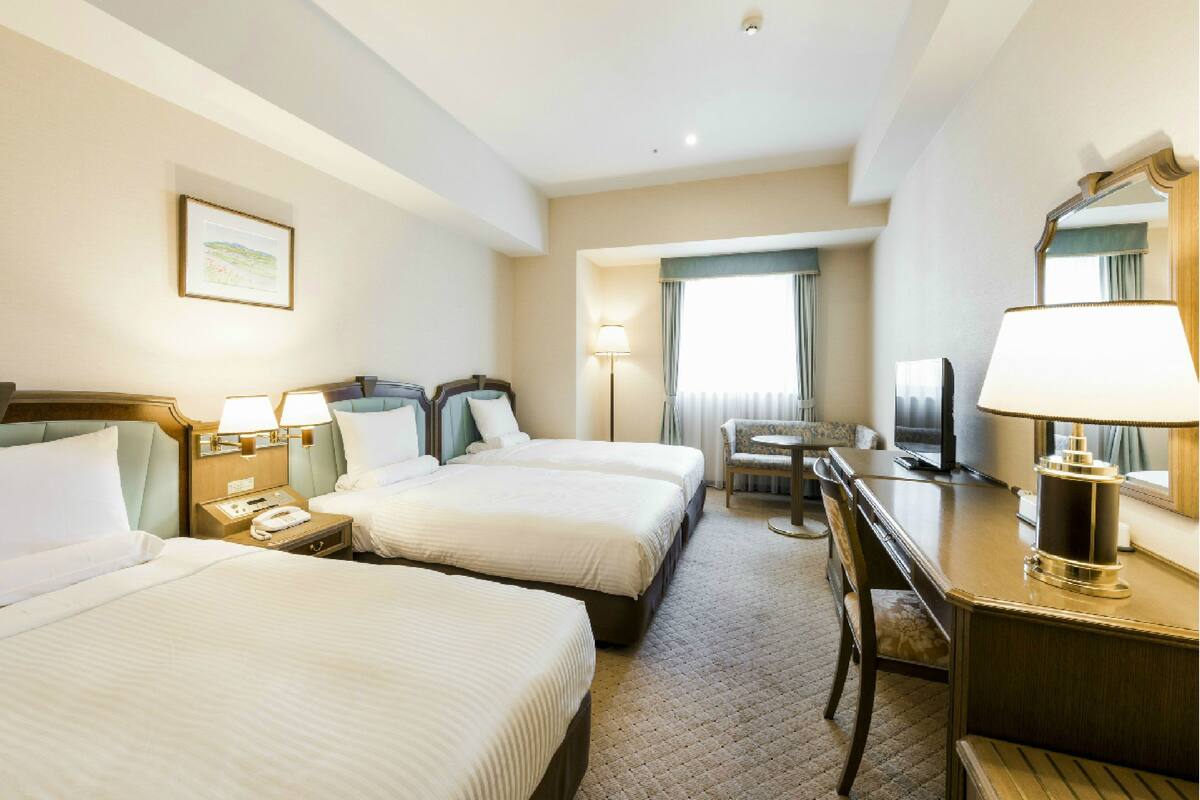 Ein Hotelzimmer mit zwei Betten, einem Schreibtisch, einem Stuhl, einem Fernseher und einem Fenster mit Vorhängen.