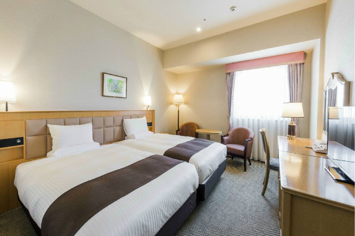 Ein Hotelzimmer mit zwei Betten, einem Schreibtisch, einem Stuhl, einer Lampe, einem Fenster mit Vorhängen und einem Bild an der Wand.