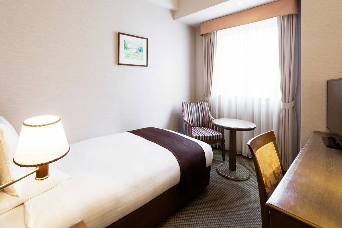 Ein Hotelzimmer mit Bett, Schreibtisch, Stuhl, Lampe, Fernseher und einem Fenster mit Vorhängen.