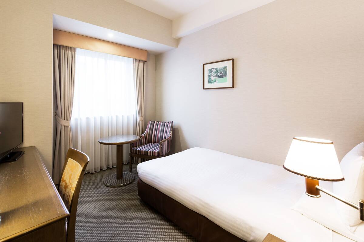 Ein Hotelzimmer mit Bett, Schreibtisch, Stuhl, Lampe, Fernseher und einem Fenster mit Vorhängen.