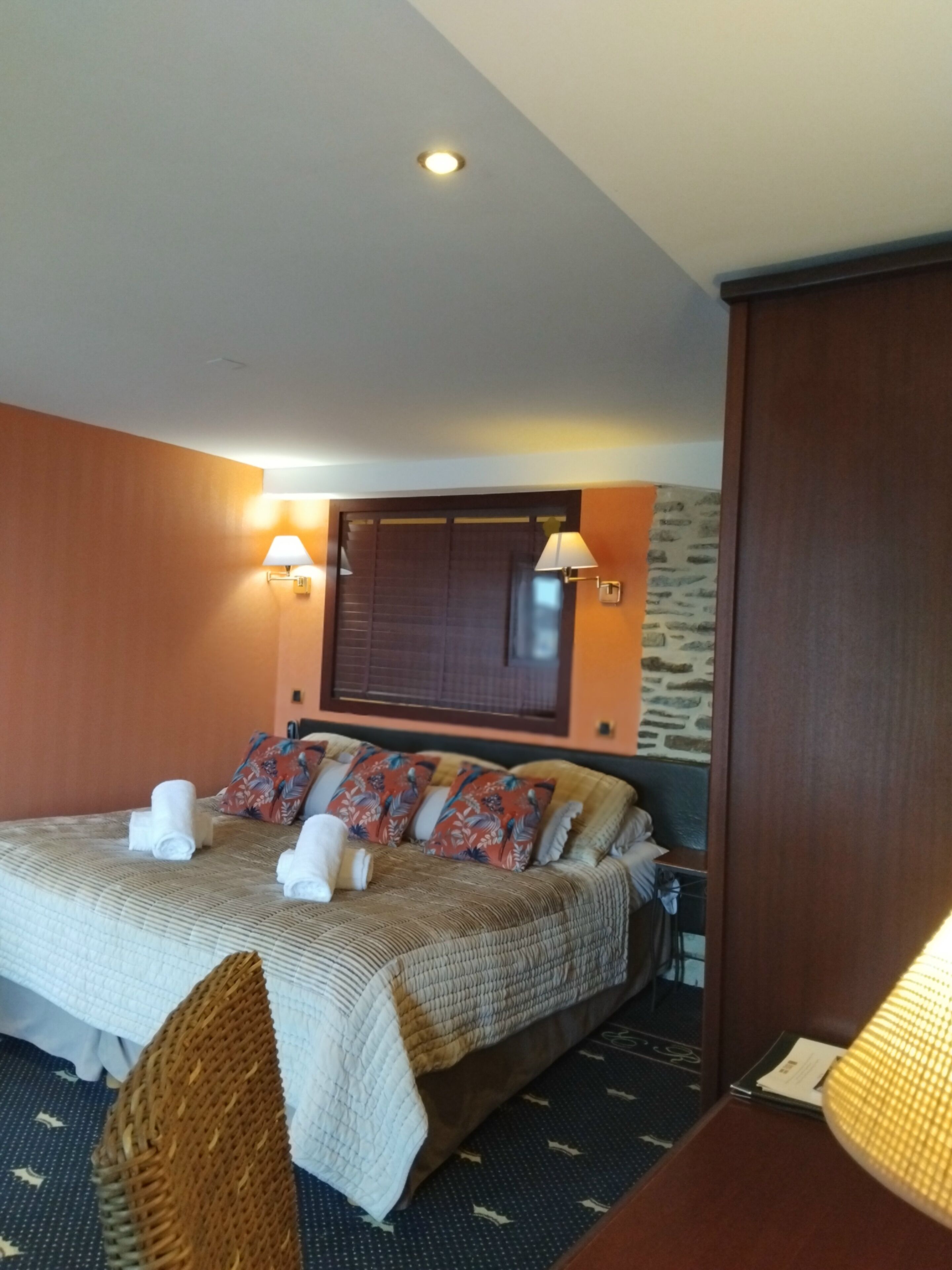 Una habitación de hotel con una cama, dos lámparas fijadas a la pared, una ventana con persianas y una pequeña mesa con lámpara.