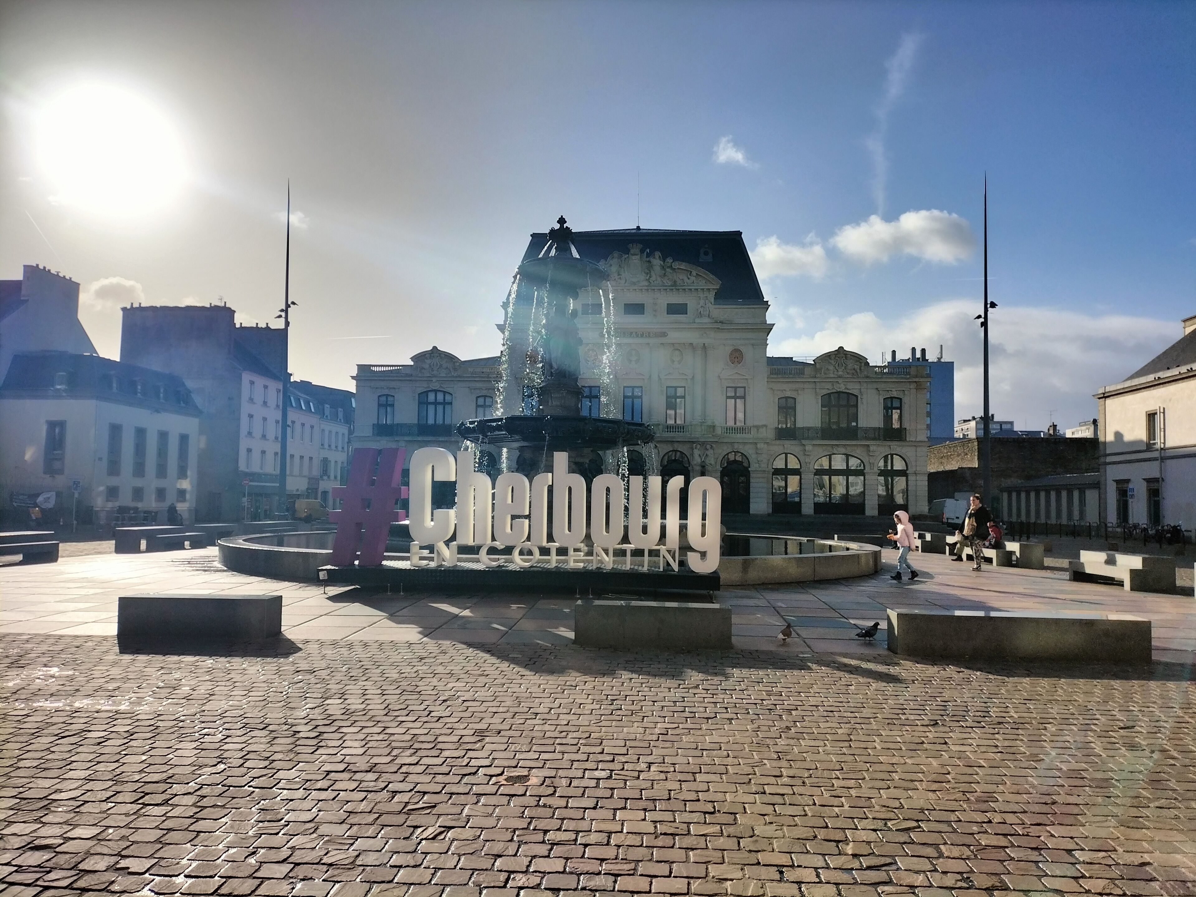 A cobblestone plaza with a fountain and a large sign reading "Cherbourg".