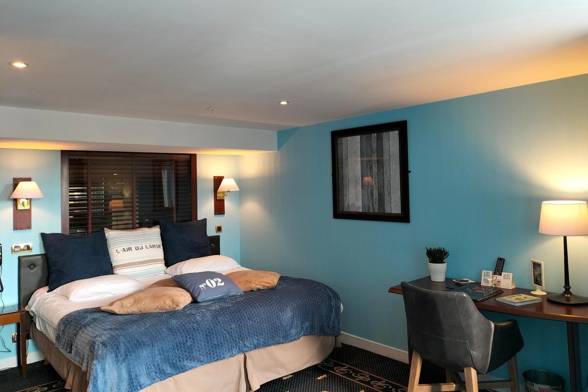 Quarto de hotel com cama, escrivaninha, cadeira, janela com persianas e luminária na parede.