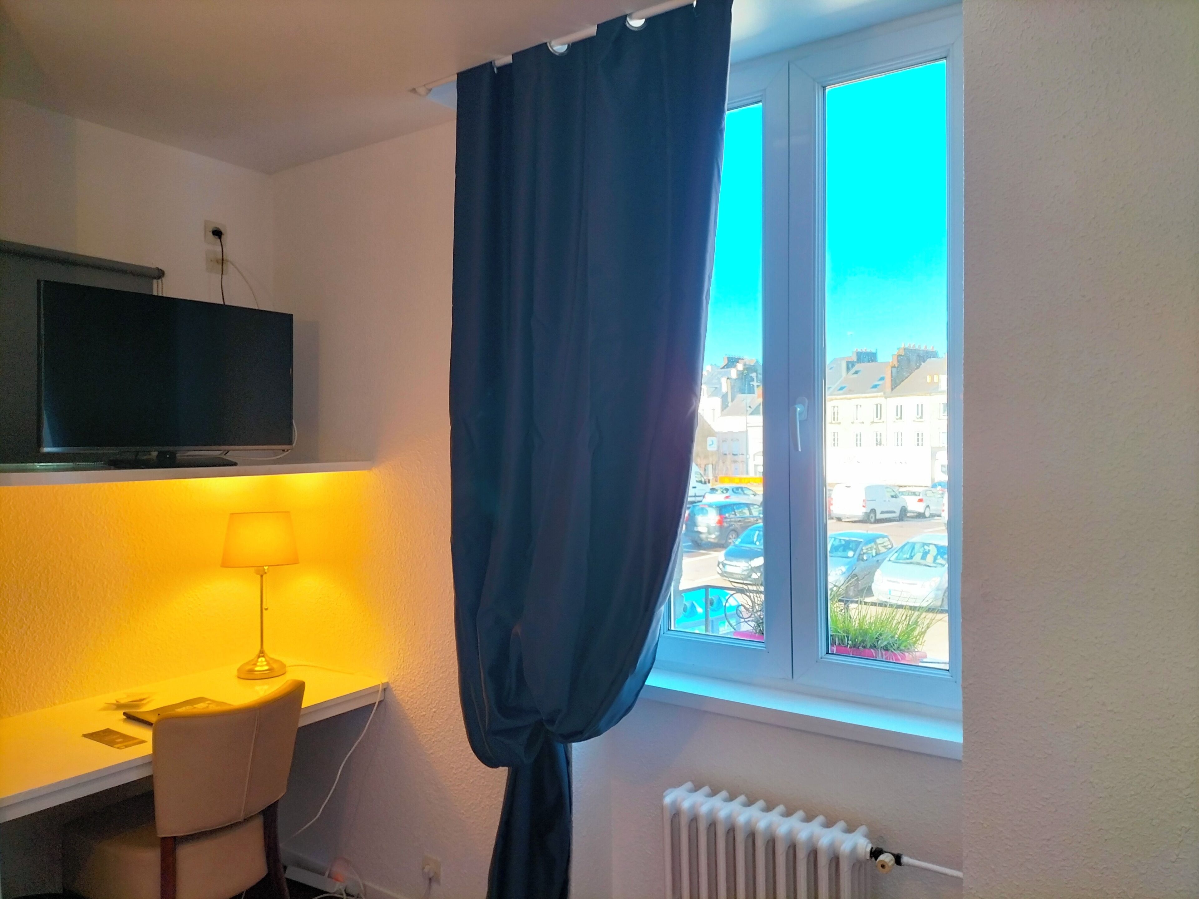 A room with a desk, chair, lamp, TV, and a window with curtains.