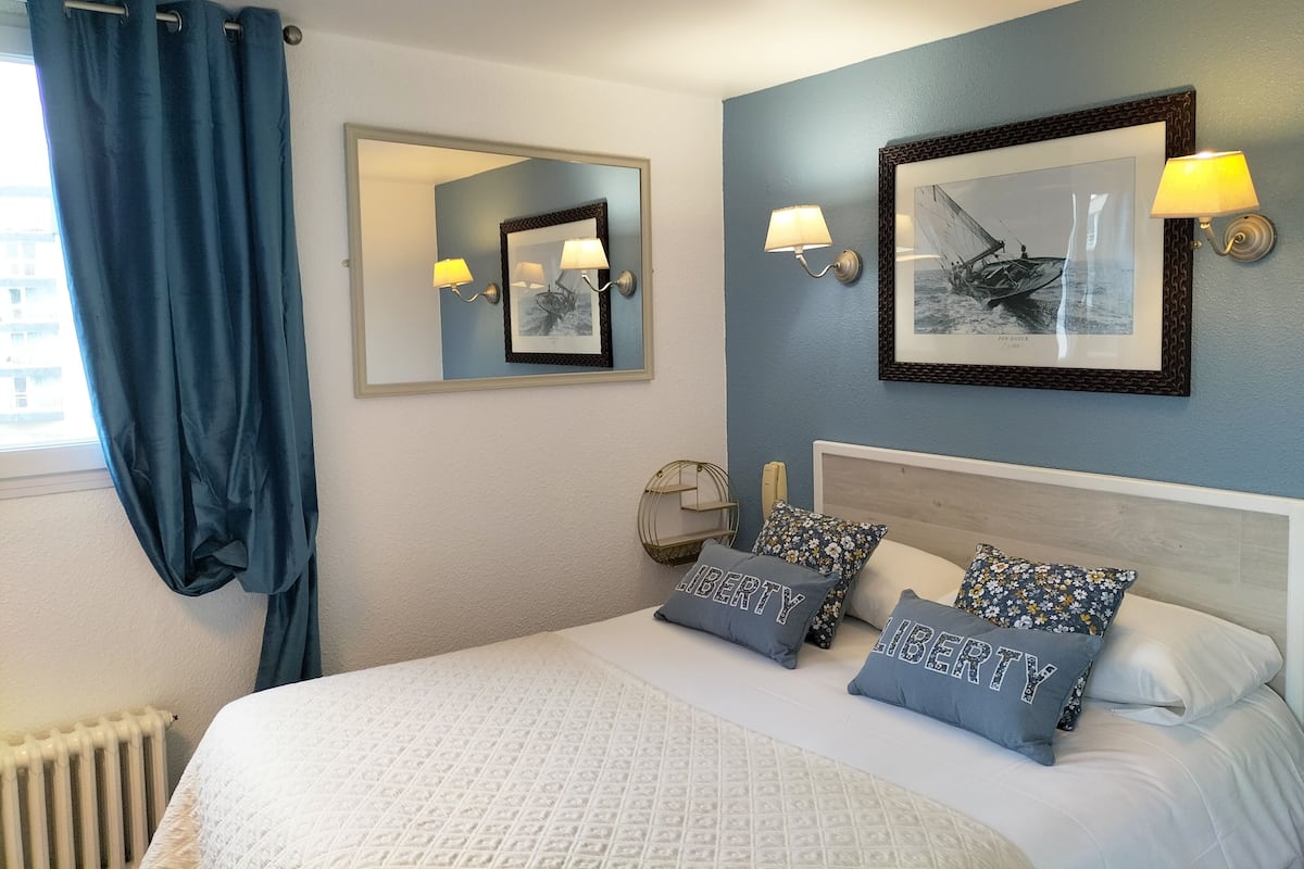 Habitación de hotel con cama, un espejo grande, dos lámparas de pared, un cuadro enmarco y una ventana con cortinas azules.