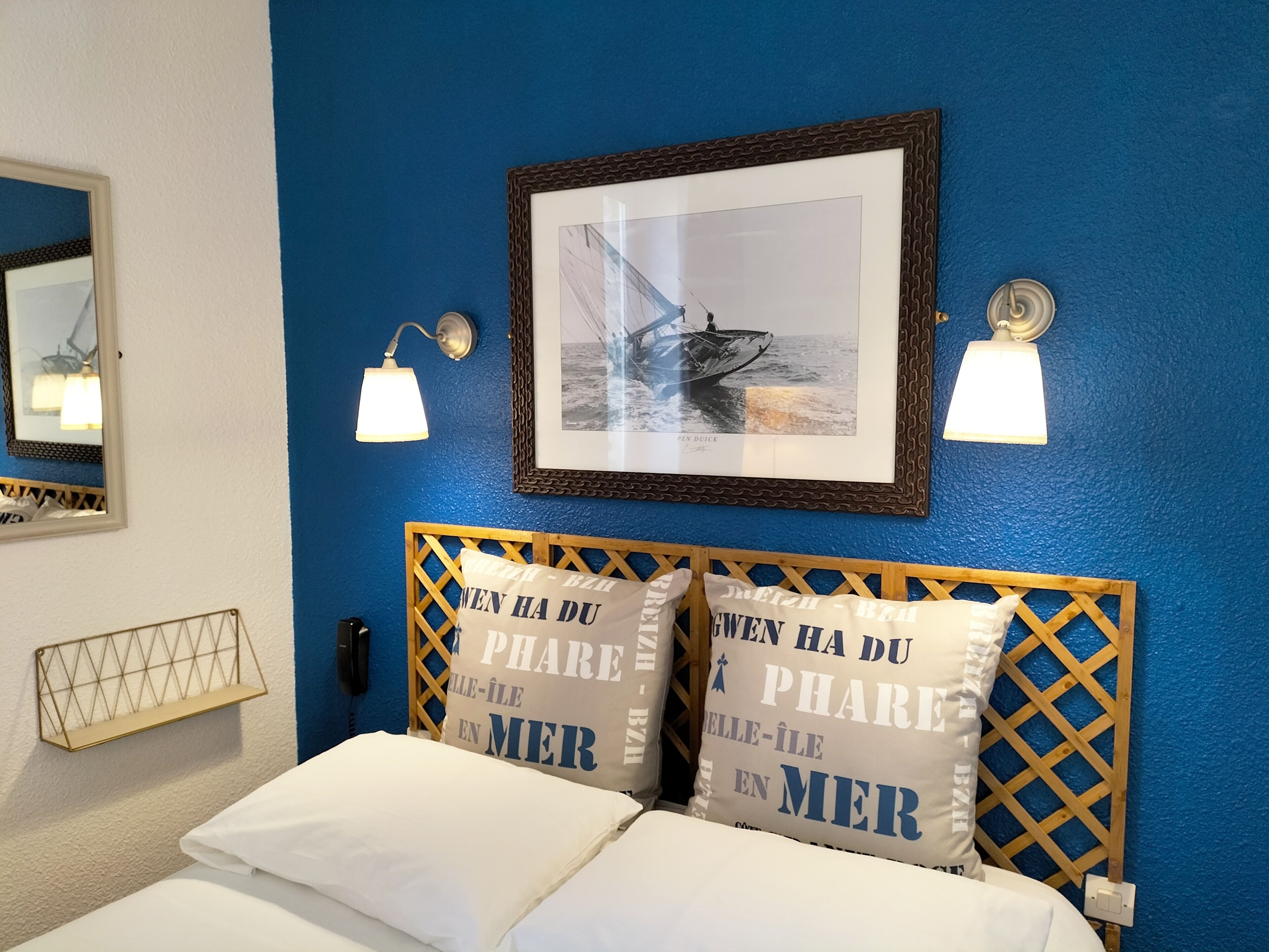 A hotel room with a blue wall, a framed photograph of a boat, two bedside lamps, and pillows with text.