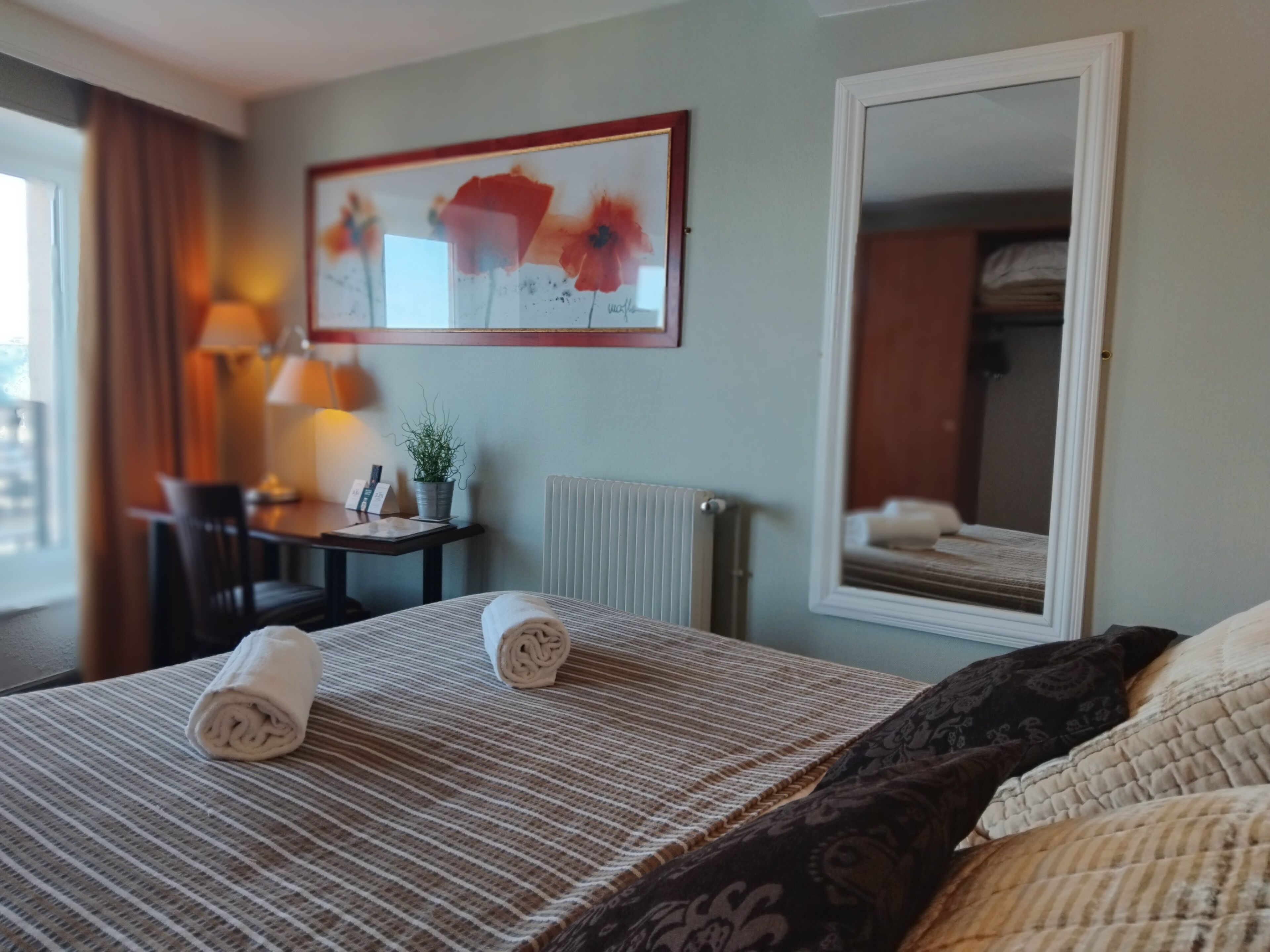A hotel room with a bed, a desk, a chair, a lamp, a framed artwork, and a mirror.