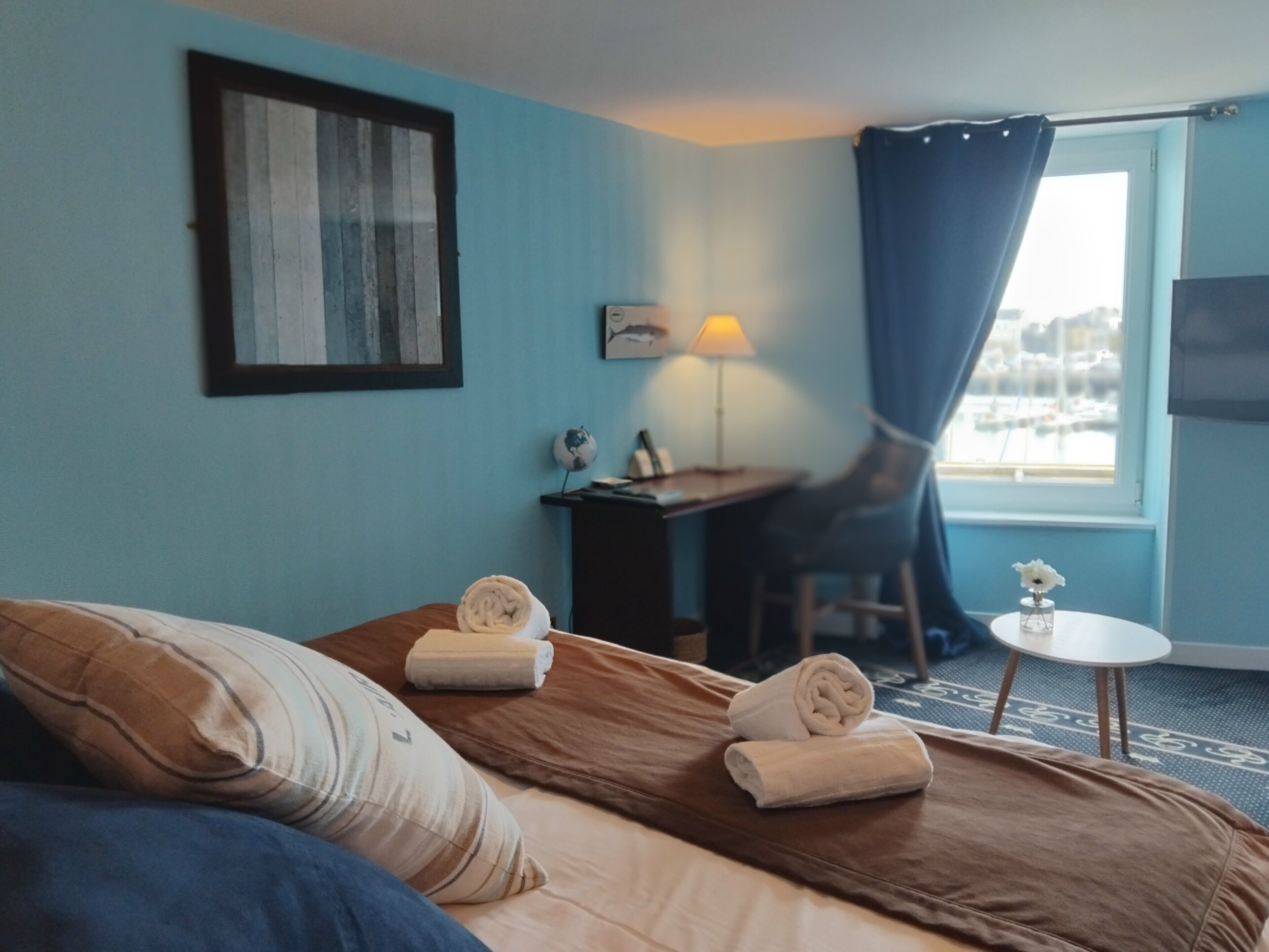 Ett hotellrum med en säng, ett skrivbord, en stol, en lampa, ett litet bord med en vas och ett fönster med gardiner.