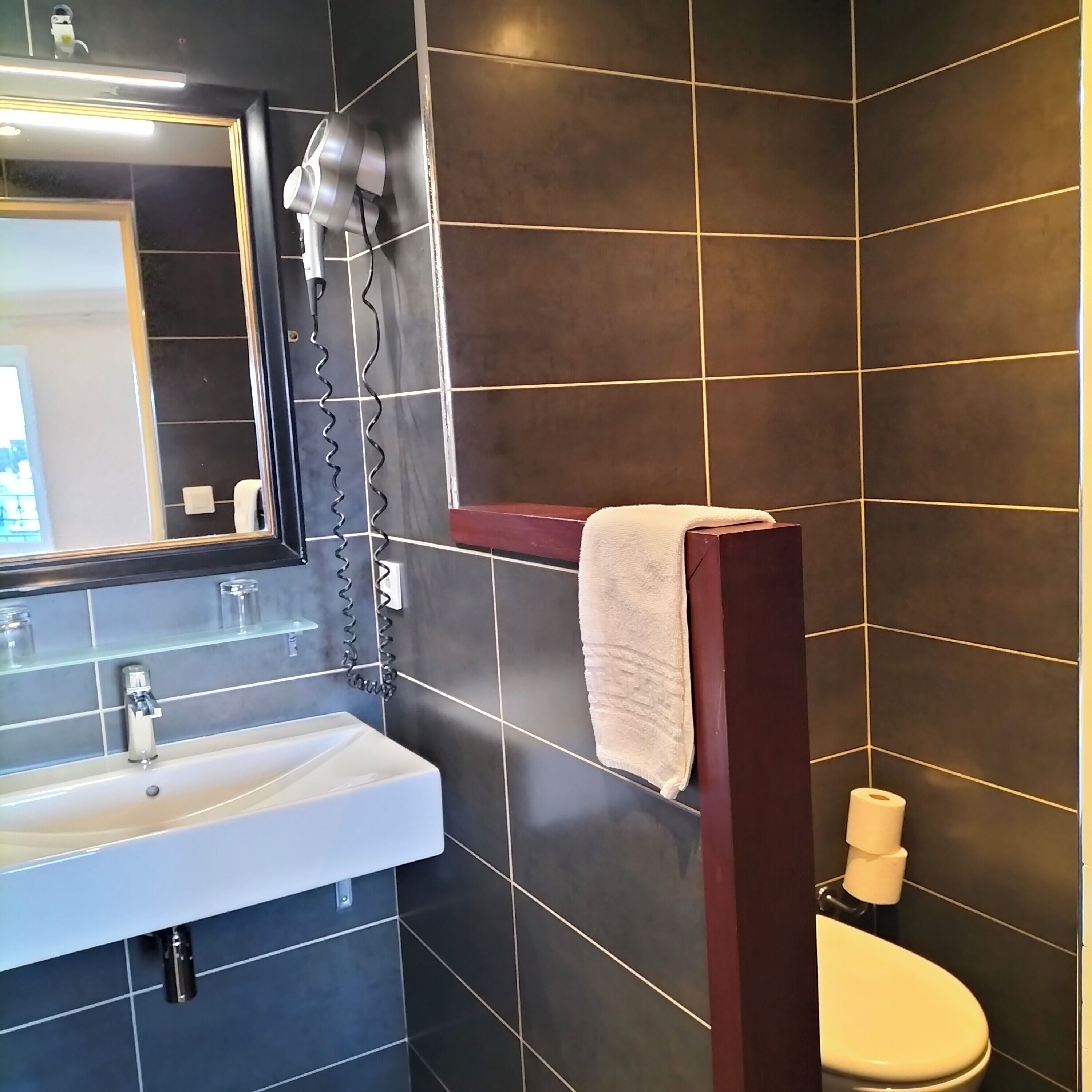 A bathroom with a white sink, dark tiled walls, a mirror, a towel rack, and a toilet.