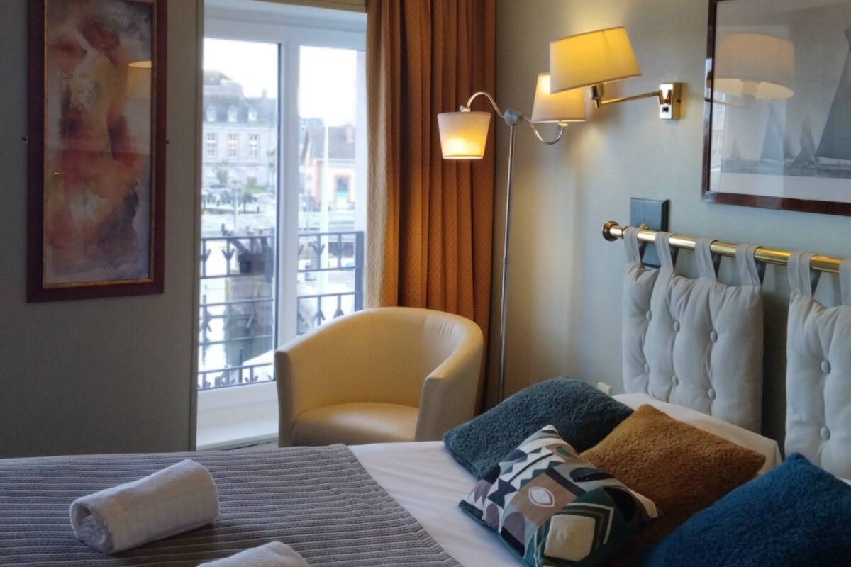 Una habitación de hotel con una cama, una silla, una lámpara de pie y un ventanal con cortinas.