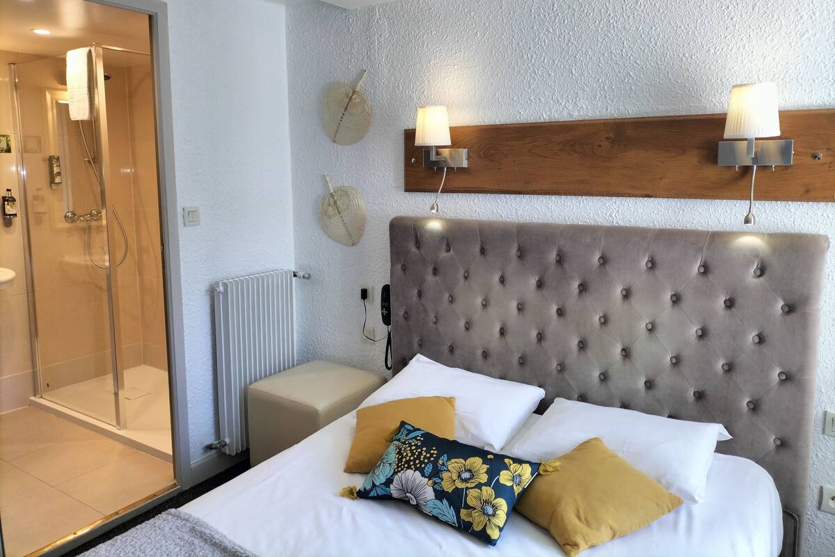 Un dormitorio con cabecera capitonada, cerramiento de ducha de cristal, mesita de noche y lámpara de pared.