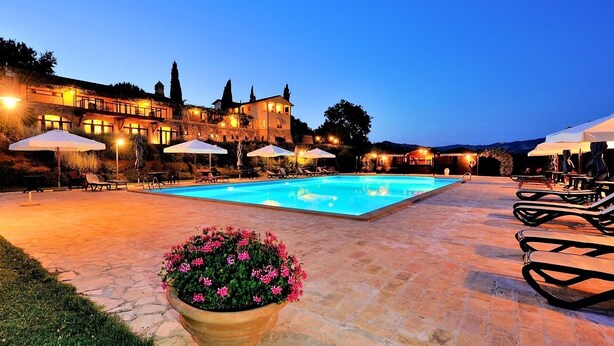 Un área de piscina con sillones de descanso y sombrillas, rodeada por un patio de piedra y un edificio grande con múltiples ventanales.