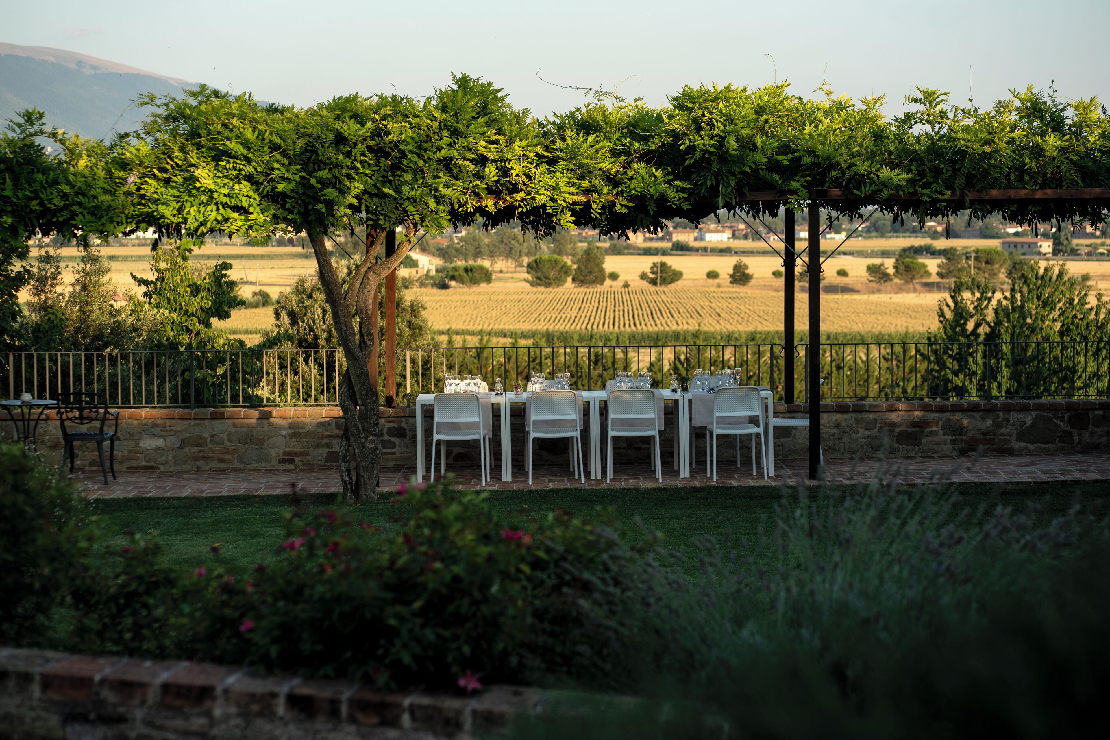 An outdoor dining area with tables and chairs under a pergola, surrounded by a garden and fields.