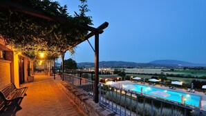 Un espacio al aire libre con pavimento de piedra, una piscina, bancos y una pérgola con plantas colgantes.