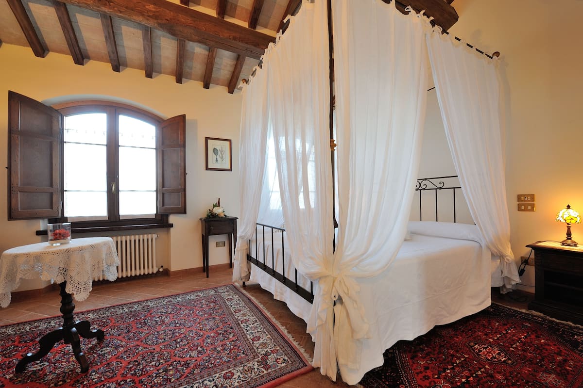 A bedroom with a four-poster bed, a large window with curtains, a small table with a lace tablecloth, and a patterned rug.