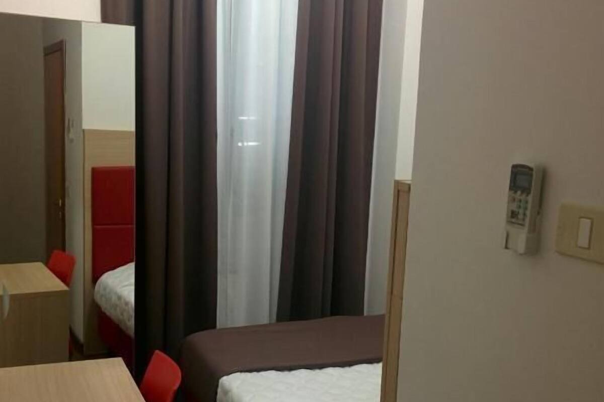 Quarto de hotel com cama, escrivaninha, cadeira, espelho, telefone e janela com cortinas.