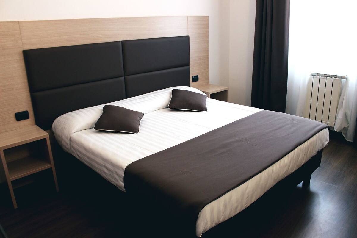 Quarto de hotel moderno com uma cama grande, duas almofadas, uma mesa de cabeceira e um criado-mudo.