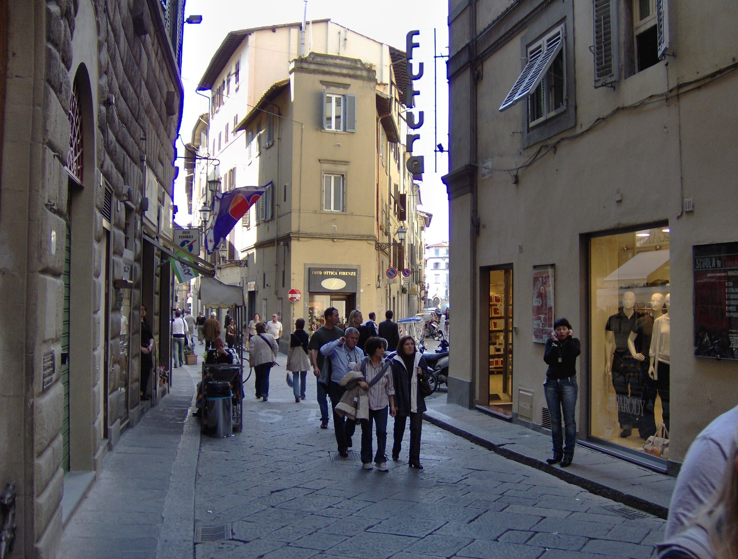 A narrow street with pedestrians, shops, and a building with a sign reading "POTTO OTICA FIRENZE".