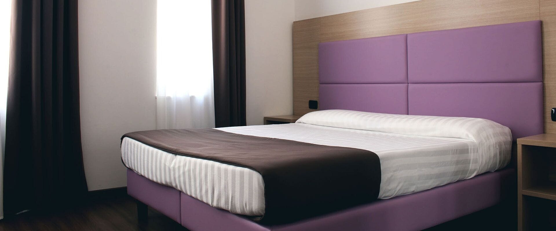 A modern hotel room with a large bed, a wooden headboard, and a nightstand.