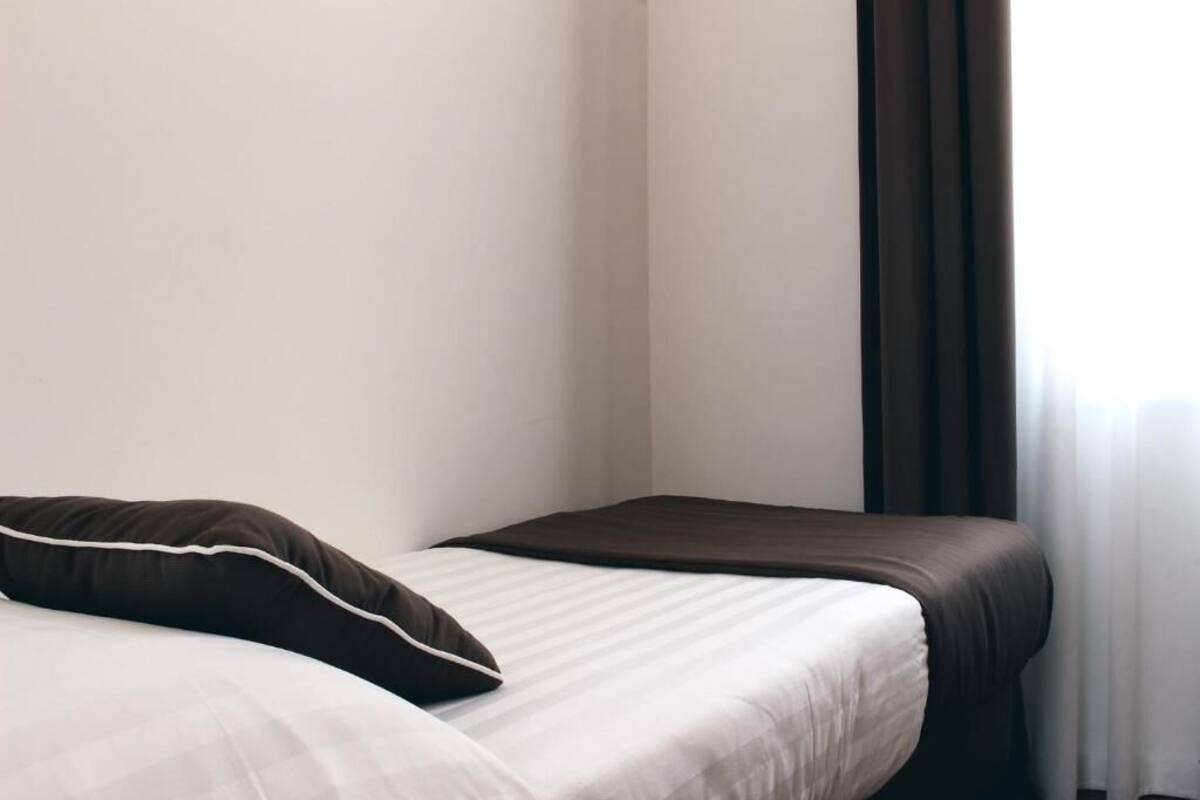 Uma cama de solteiro com um espalha cama branco e castanho, um travesseiro marrom escuro e uma janela com cortinas escuras.