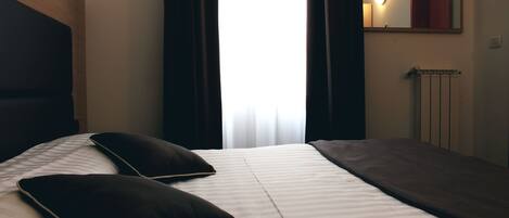 A hotel room with a bed, a headboard, a window with curtains, a mirror, and a radiator.