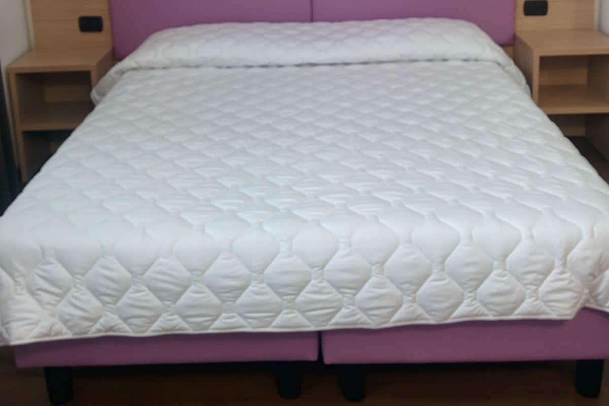 Cama com cabeceira de madeira e detalhe roxo, com duas mesas de cabeceira de cada lado.
